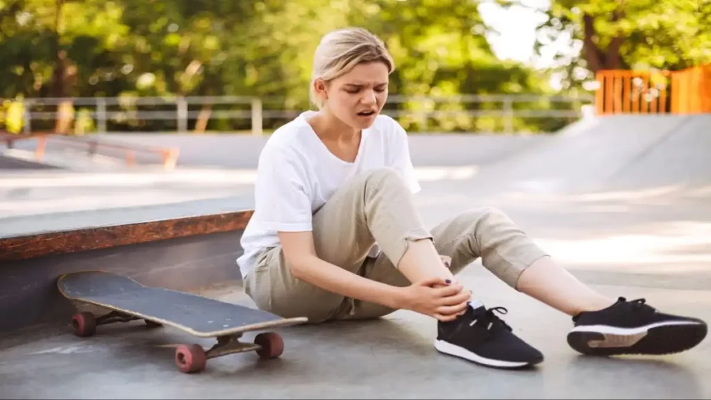 How Your Shoes Impact Your Knee Pain. A girl wearing a white shirt, with her skateboard, sits on the road while holding her knee in pain